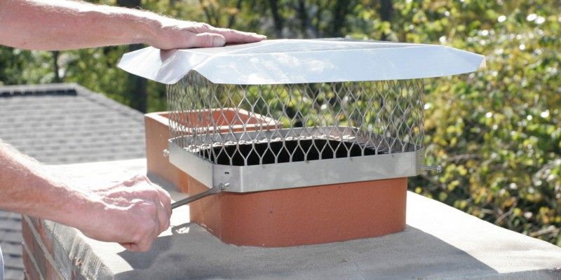 A man using a screwdriver to install a square stainless steel chimney cap on a flue tile.