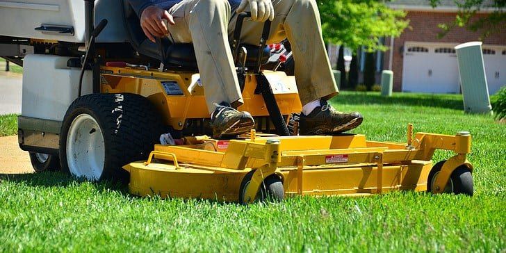 An image showing a man from the waist down sitting on a yellow zero turn lawn mower on a suburban lawn