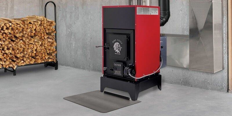 A Fire Chief FC1000E wood burning furnace in a basement. To the left is a firewood rack with logs, and in front of the furnace is a steel stove board.