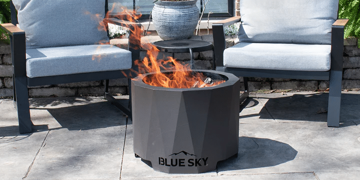 A Blue Sky Improved Peak Smokeless Patio Fire Pit sitting ablaze on a concrete patio with two gray chairs in the background and a brick wall behind them