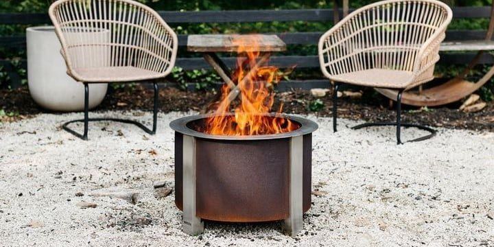 A Breeo X19 smokeless fire pit sitting ablaze on white sand with two beige patio chairs in the background and a gray wooden fence behind them