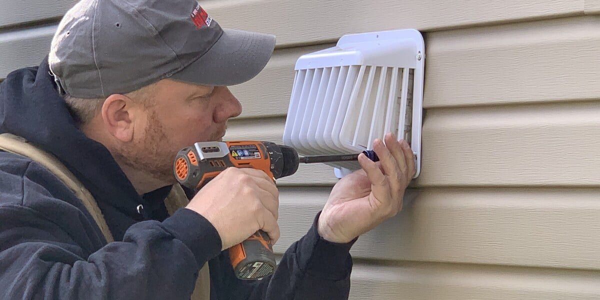 A man with a gray hat installing a white, plastic dryer vent guard on a house with beige siding using an orange drill.