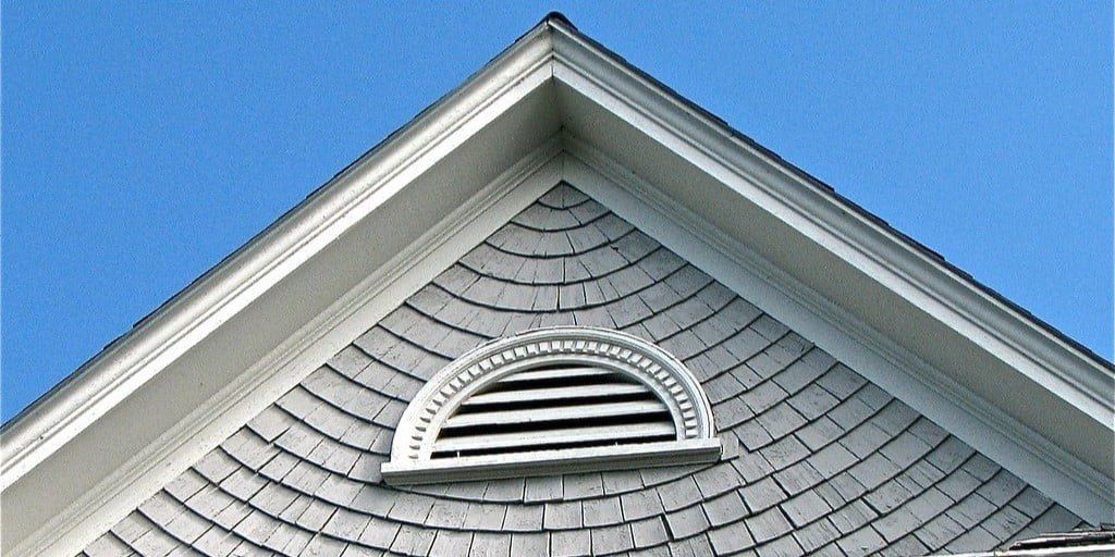 A half-circle gable vent against gray siding and a pointed roof with a blue sky in the background