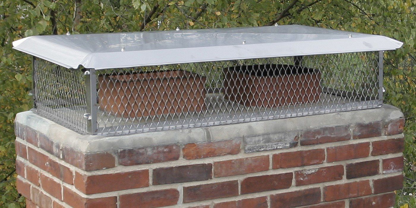A stainless steel multi-flue chimney cap installed on a chimney crown with foliage in the background