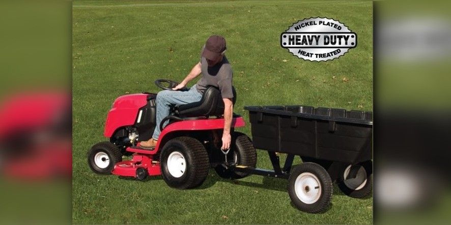 A man in jeans, a black shirt, and a black hat on a red lawn tractor reaching back to pull out a Good Vibrations Quick Connect Hitch Pin.