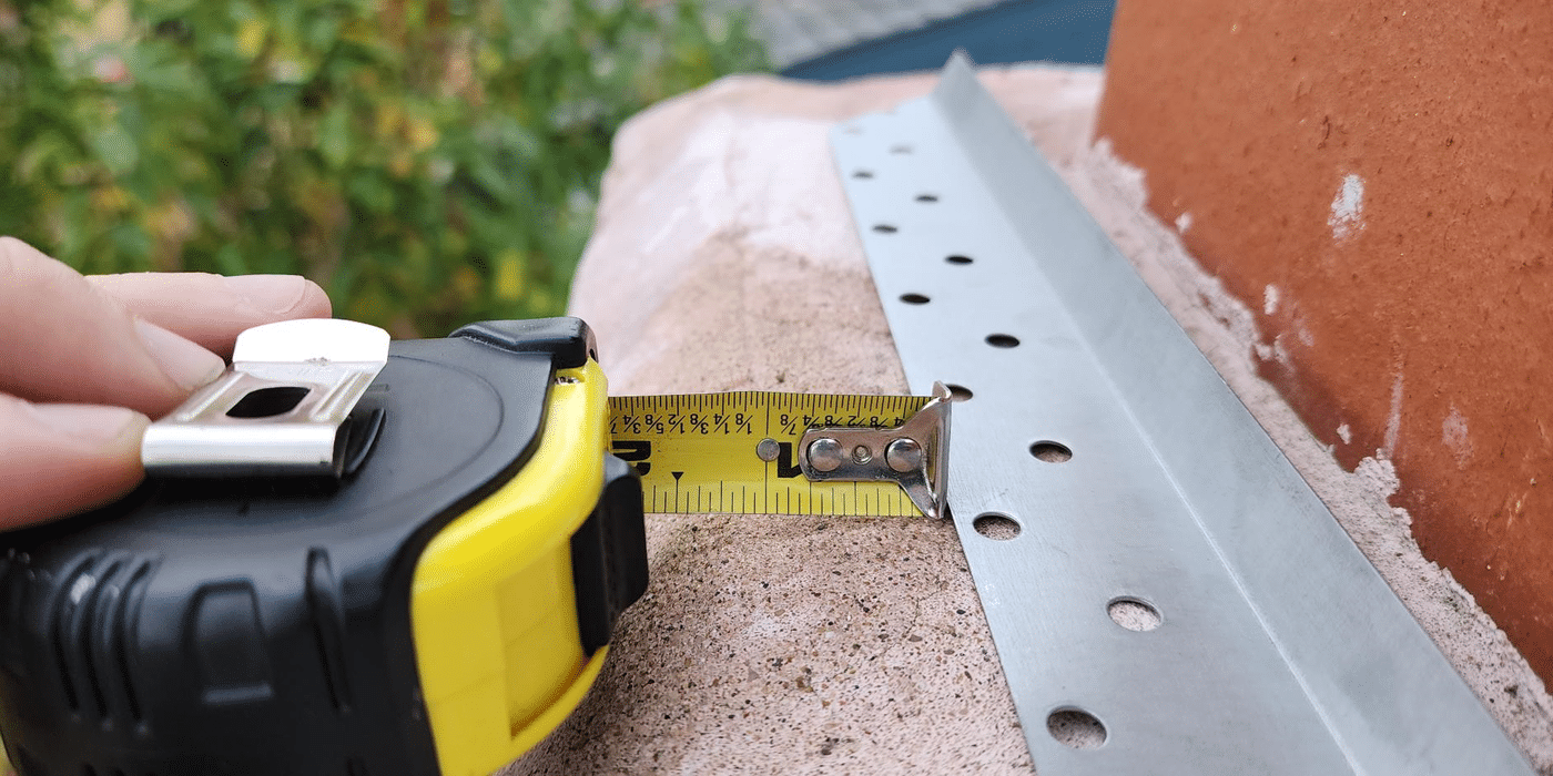 A multi-flue chimney cap flange resting on a chimney crown with a tape measure indicating that the flange is at least one inch from the edge of the crown