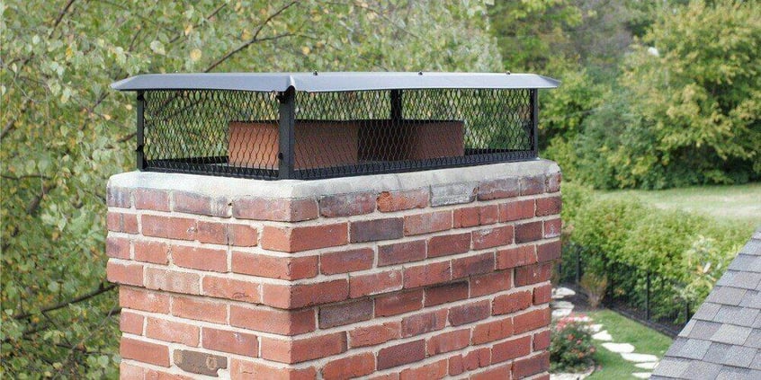 A Draft King Big Top Black Galvanized Steel Multi-Flue Chimney Cap installed on a multi-flue chimney. The home's yard can be seen in the background.