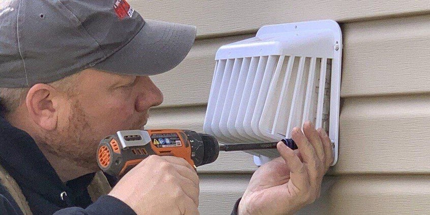 A man installing a plastic, bolt-on dryer vent cover on the side of a house with beige siding. The man is using an orange drill.