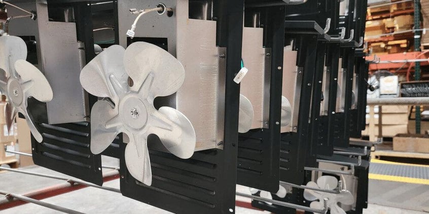 A factory rack holding the backs of multiple Magic Heat heat reclaimers. On the front-most panel, a five-prong fan is visible.