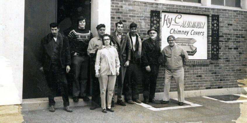 A black-and-white photo of HY-C employees standing outside of the factory in front of a HY-C Aluminum Chimney Covers sign circa 1960.