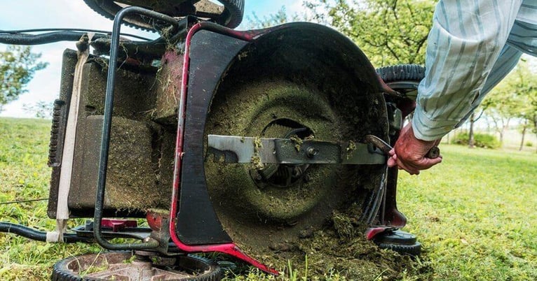 A lawnmower lying on its side on a lawn as a man uses a tool to scrape grass from the mower blades and deck.