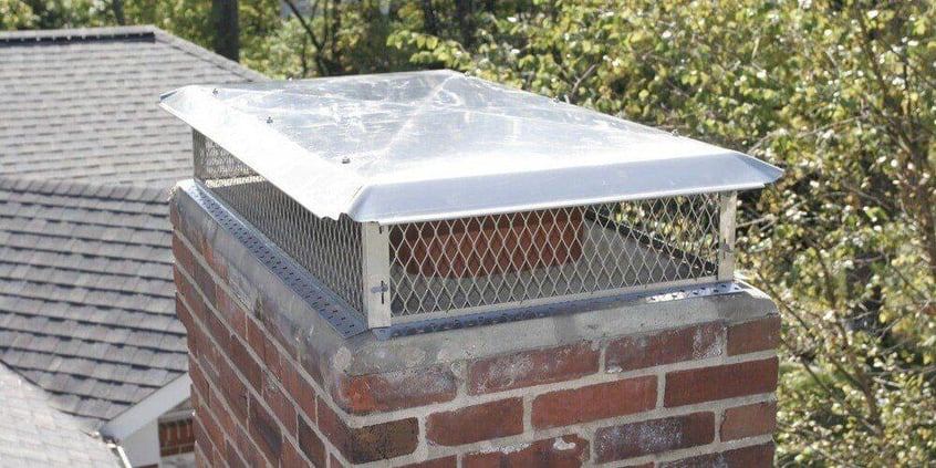 A stainless steel Draft King Big Top Multi Flue Chimney Cap installed on a masonry chimney. A roof and trees are visible in the background.