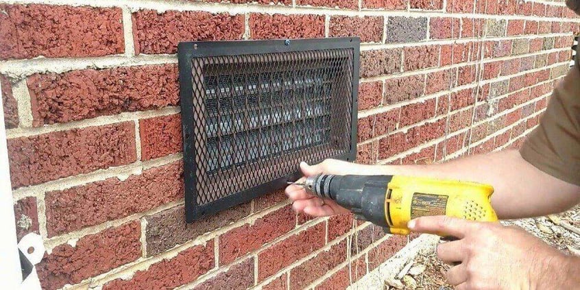 A man using a yellow drill to install a HY-GUARD EXCLUSION Foundation Vent Guard over a foundation vent on a brick wall.