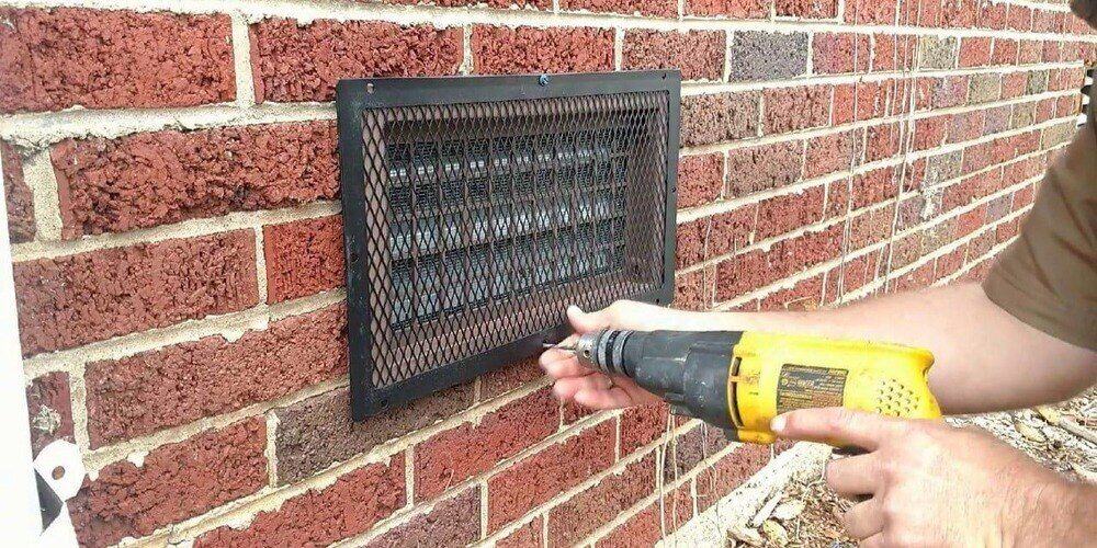 A man using a yellow drill to install a HY-GUARD EXCLUSION Foundation Vent Guard over a foundation vent on a brick wall.