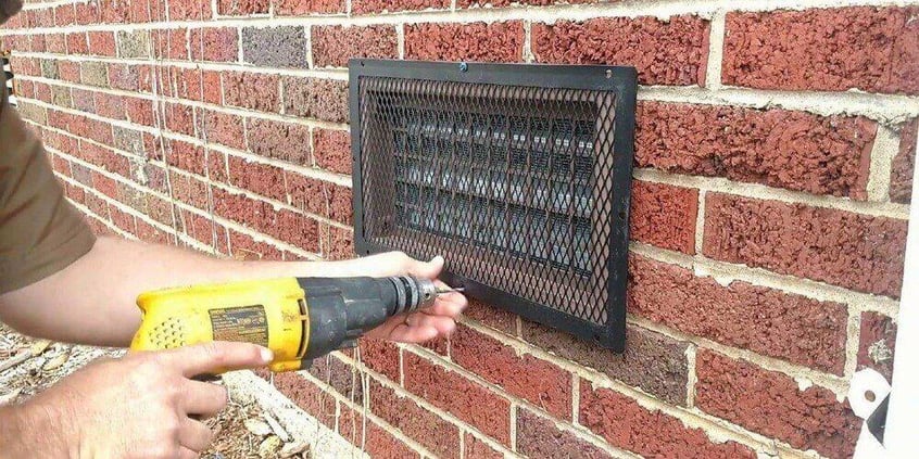 A man using a yellow drill to install a HY-GUARD EXCLUSION Foundation Vent Guard over a foundation vent on a brick wall.
