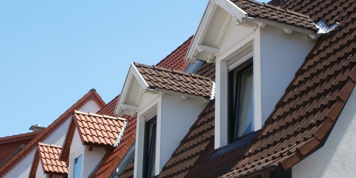 A steep roof with dark brown terracotta shingles with two white dormers sticking out against the background of a blue sky