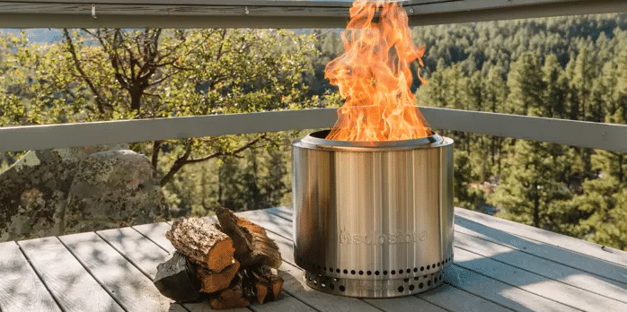 A Solo Stove Bonfire 2.0 sitting ablaze on the corner of a wooden patio with a forest in the background and wood fuel beside it