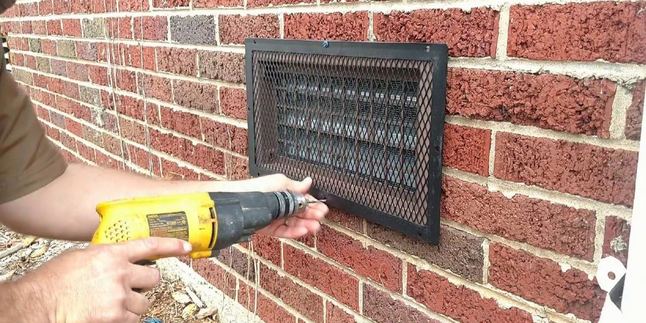 HY-GUARD EXCLUSION foundation vent guard installed on the side of a brick house by an installer using a yellow drill