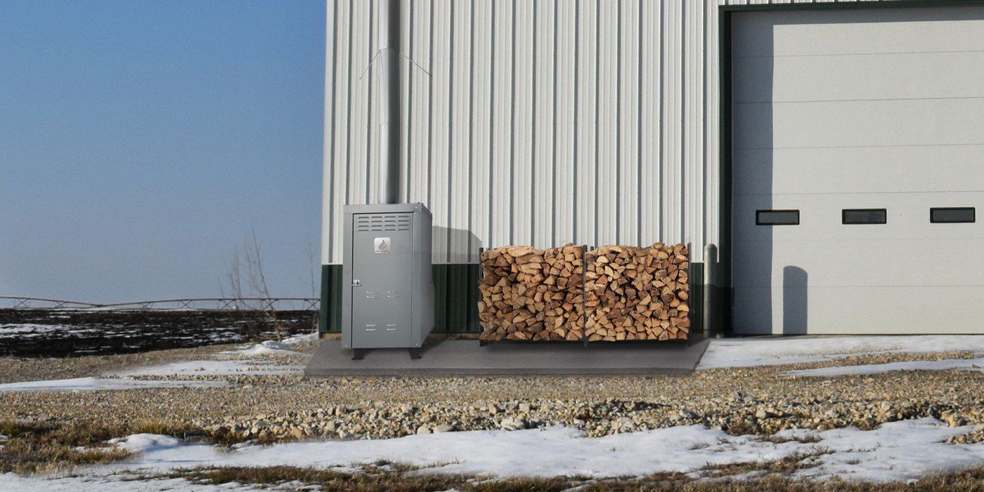 An outdoor Ignite Tech furnace sitting on a concrete slab outside of a commercial building with a cord of wood sitting next to it
