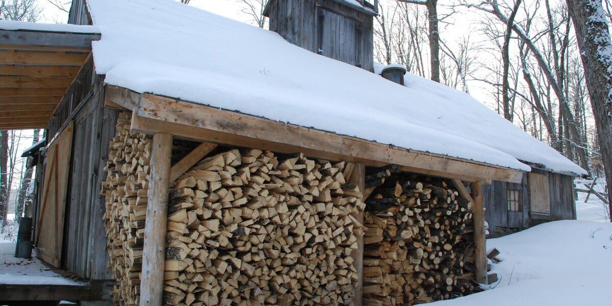 A wood log rack stuffed with over a cord of wood in front of a wood cabin covered in snow in the middle of the woods