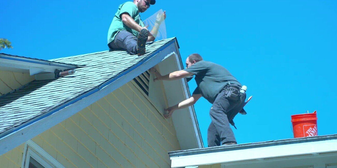 Two men on a roof installing triangular-shaped HY-GUARD EXCLUSION Pest Armor Tight Mesh Panel over a triangular gable end vent.