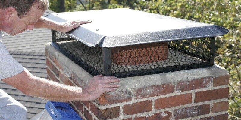 A man installing a Draft King BigTop Galvanized Steel Multi-Flue Chimney Cap on a chimney. He is on a ladder, and tree branches are visible.