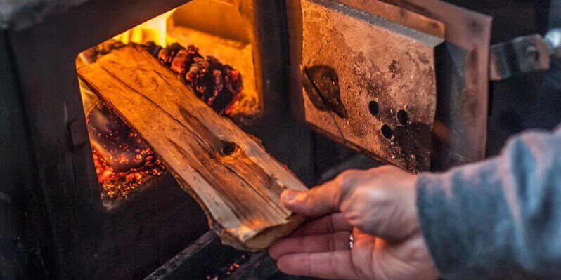 A person adding a piece of wood into a wood stove. The fire in the stove is just beginning to kindle.