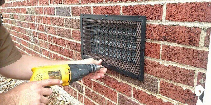 A man installing a black HY-GUARD EXCLUSION Foundation Vent Screen over a foundation vent. He is using a yellow drill to install it.