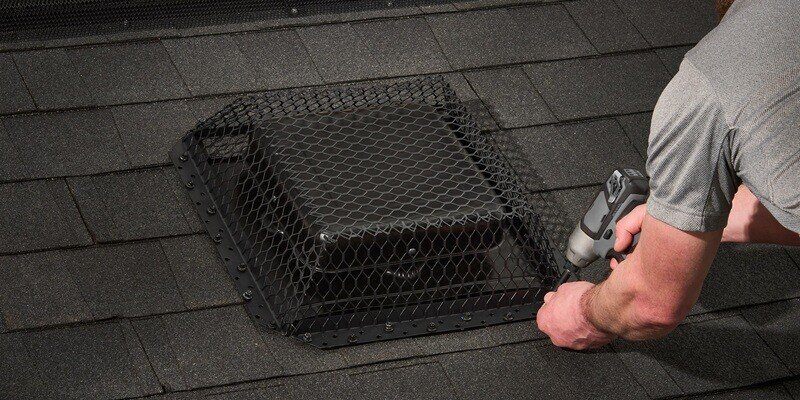 A man using a gray drill to install a HY-GUARD EXCLUSION roof vent cover over a roof vent on a roof with black shingles.