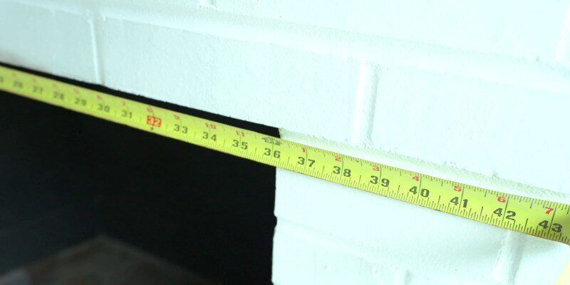 A measuring tape across the top of a white-bricked fireplace opening. The tape shows that the fireplace opening measures 36 inches.
