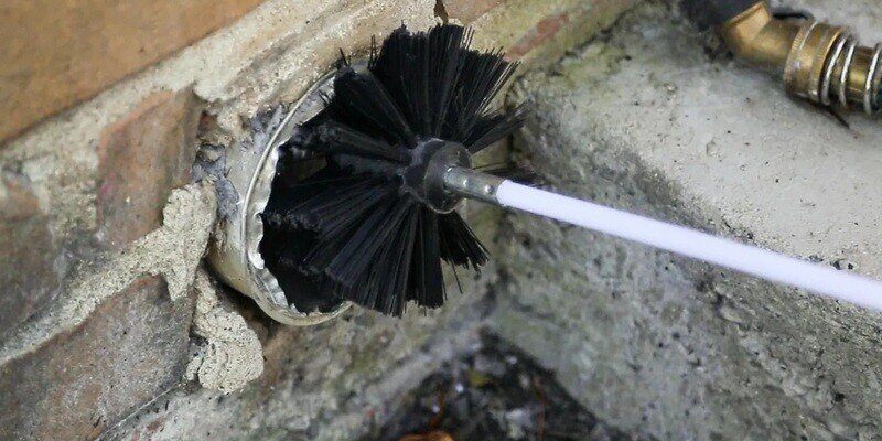 A dryer vent cleaning kit's brush head entering a dryer vent from the outside of a home. The wall that houses the vent is made of brick.
