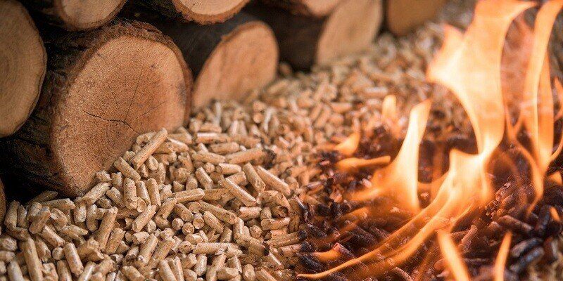 A closeup of a pile of wood pellets next to a pile of round logs. On the right side, the wood pellets are on fire.