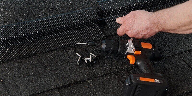 A man installing HY-GUARD EXCLUSION Pest Armor on a roof deck vent. The installation hardware and a black and orange drill rest on the shingles.