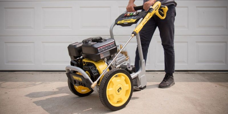A man pushing a large, heavy pressure washer on his driveway in front of a garage door.
