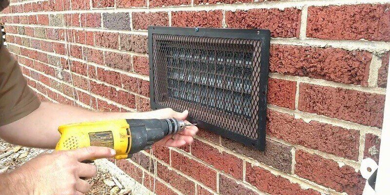 A man using a yellow drill to install a HY-GUARD EXCLUSION foundation vent cover.