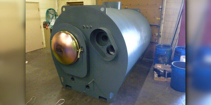 A GARN wood boiler unit sitting on a factory floor. A closed garage door is behind the unit, and blue buckets sit to its right.
