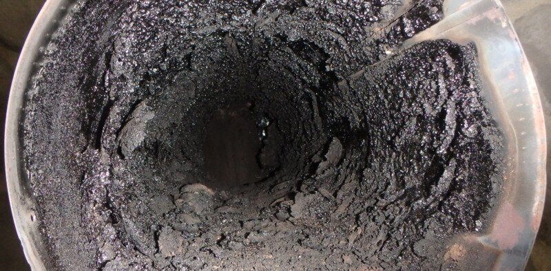 A top-down view of a section of round metal chimney pipe. A thick layer of creosote has developed on the walls of the pipe.