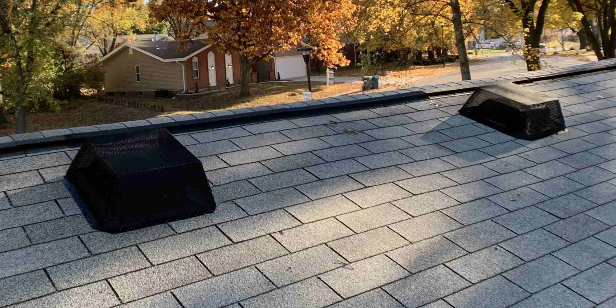 Two galvanized steel roof vent guards installed on a suburban roof over two black box vents. The rest of the neighborhood can be seen in the background.