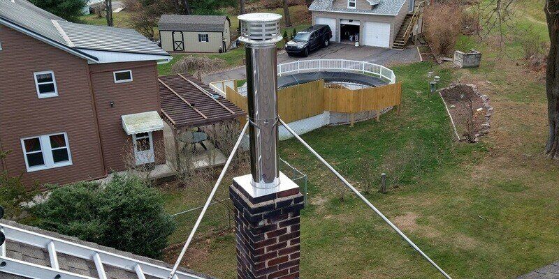 A wood burning appliance chimney protruding from a roof with a neighboring backyard visible. The chimney has a cap on it.