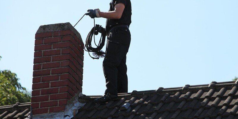 A chimney sweep standing on a roof with black terracotta shingles feeding a chimney sweep's brush into the chimney.