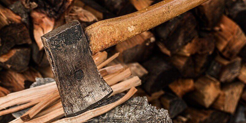 A closeup of an ax wedged into a tree stump. There is stacked firewood in the background, and kindling lying next to the ax head.