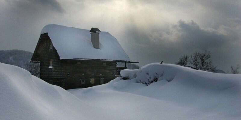 An isolated cabin among large snowdrifts. There is snow on the roof of the cabin. The sky is cloudy, but the sun is poking through a bit.