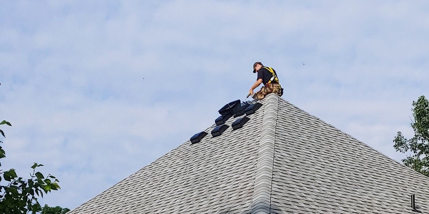 A man kneeling on the peak of a rood installing HY-GUARD EXCLUSION Roof VentGuards against the backdrop of a partly cloudy sky.