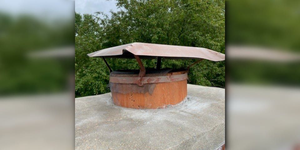 An old, bent, rusted chimney cap installed on a round flue tile. There are trees visible in the background.