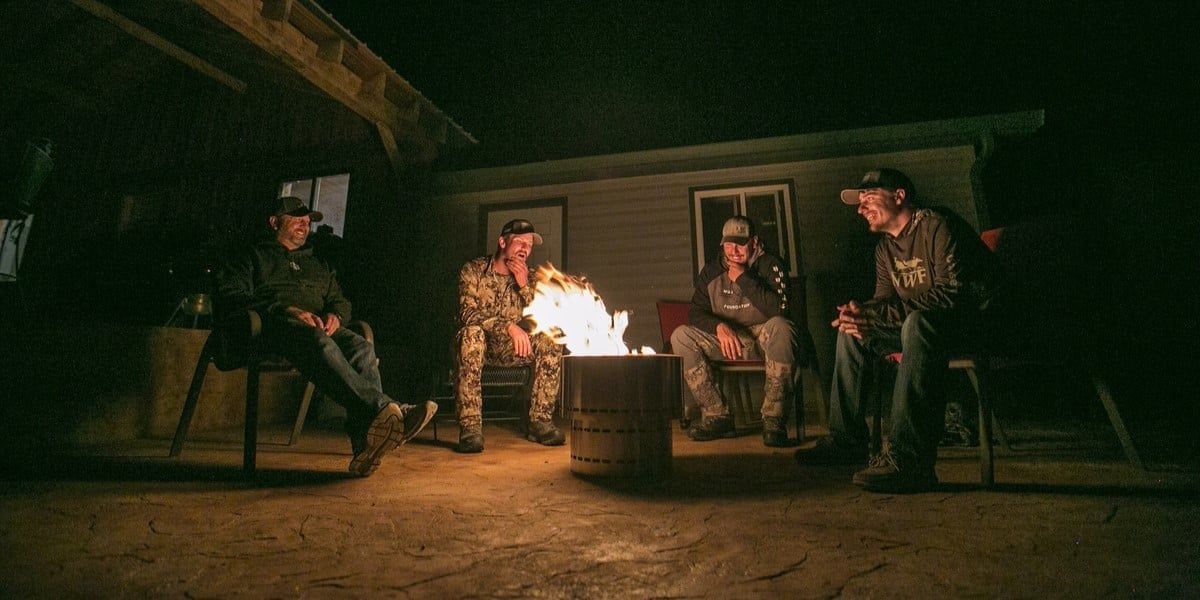 Four men sitting around a Flame Genie smokeless fire pit on a patio at night. There is a fire burning in the fire pit.