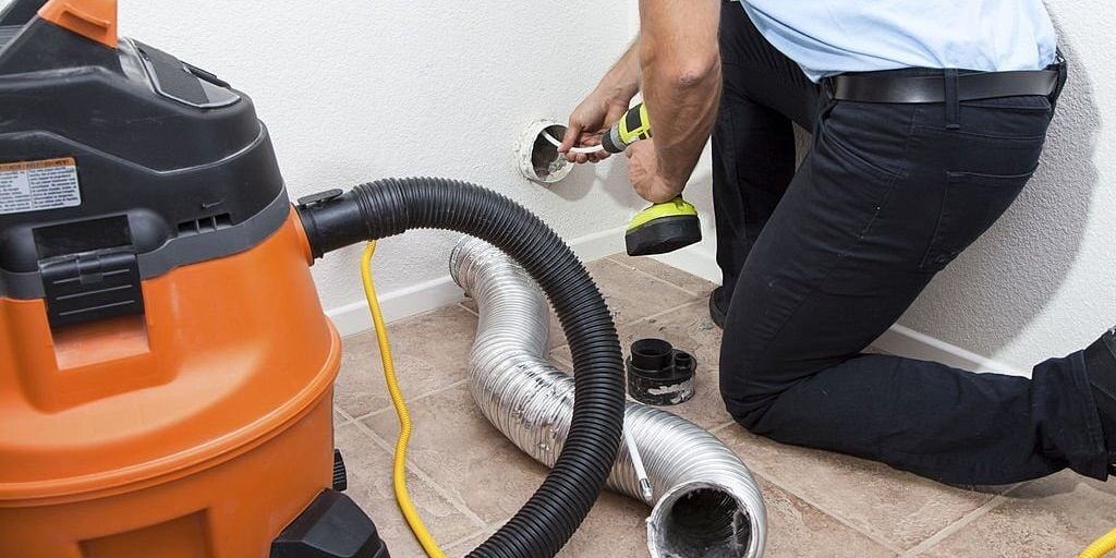 A man kneeling next to an exposed dryer vent. He's using a rotary dryer vent cleaning tool to clean the vent. There is an orange ShopVac in the foreground, and a flexible dryer vent pipe on the floor.