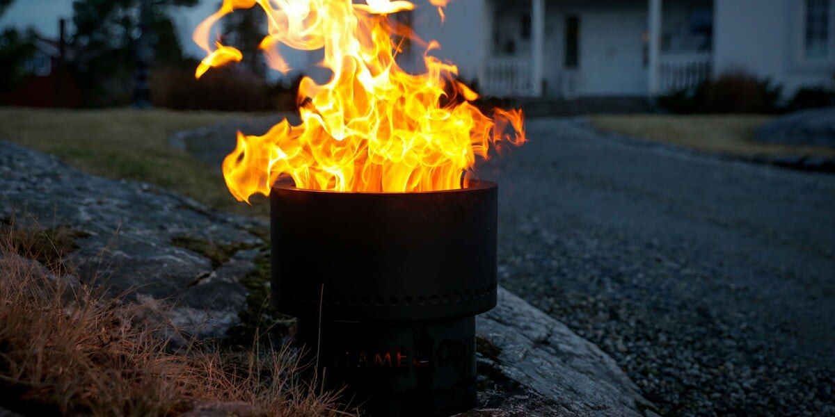 A Flame Genie smokeless fire pit sitting on the ground with a house in the background. A fire is blazing within the firepit.