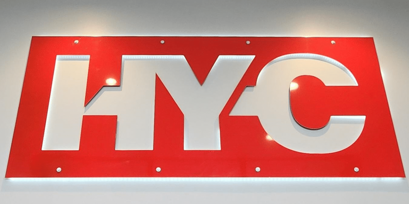 A fabricated metal HY-C logo hanging on a wall illuminated by vanity lighting.