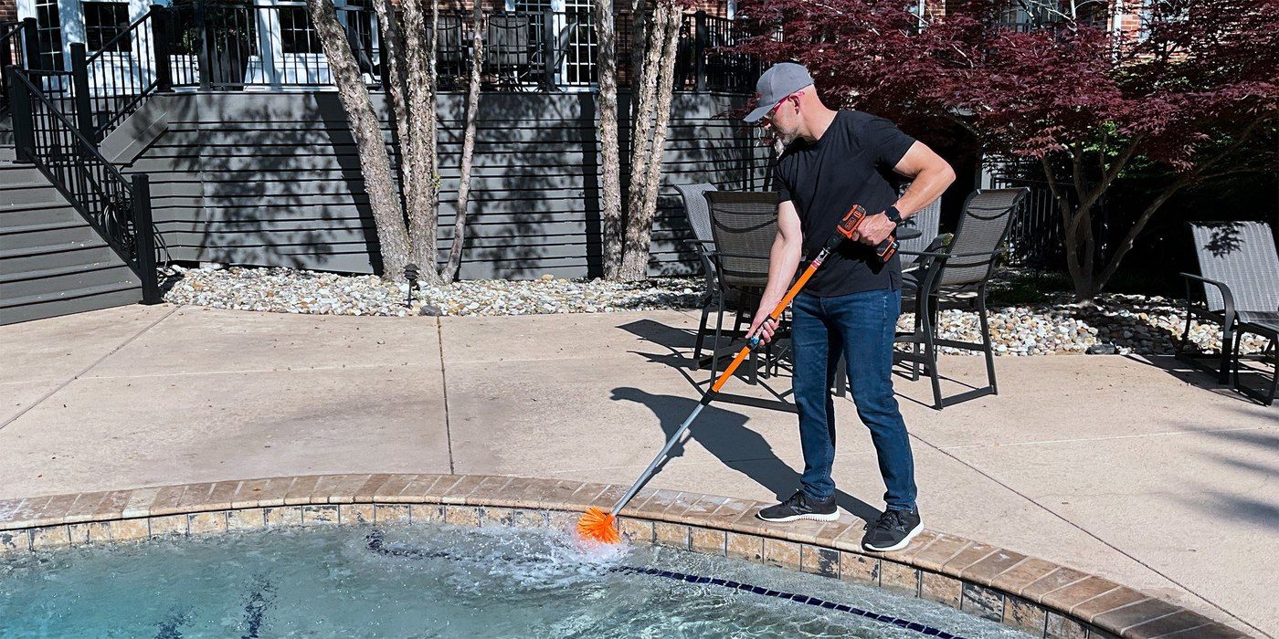 A man in blue jeans, a black t-shirt, and a gray hat using a SpinAway drill brush to clean his pool.