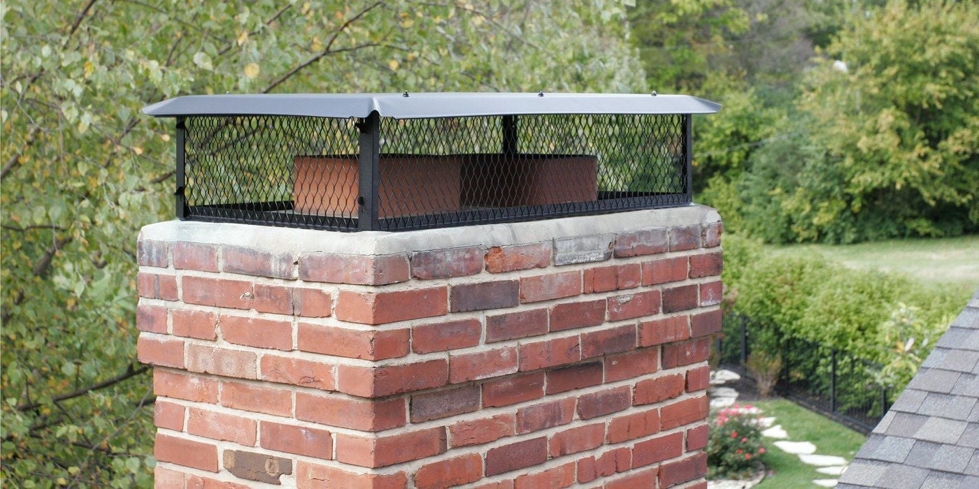 A black galvanized steel multi-flue chimney cap installed on a chimney crown with a nicely landscaped yard visible in the background.